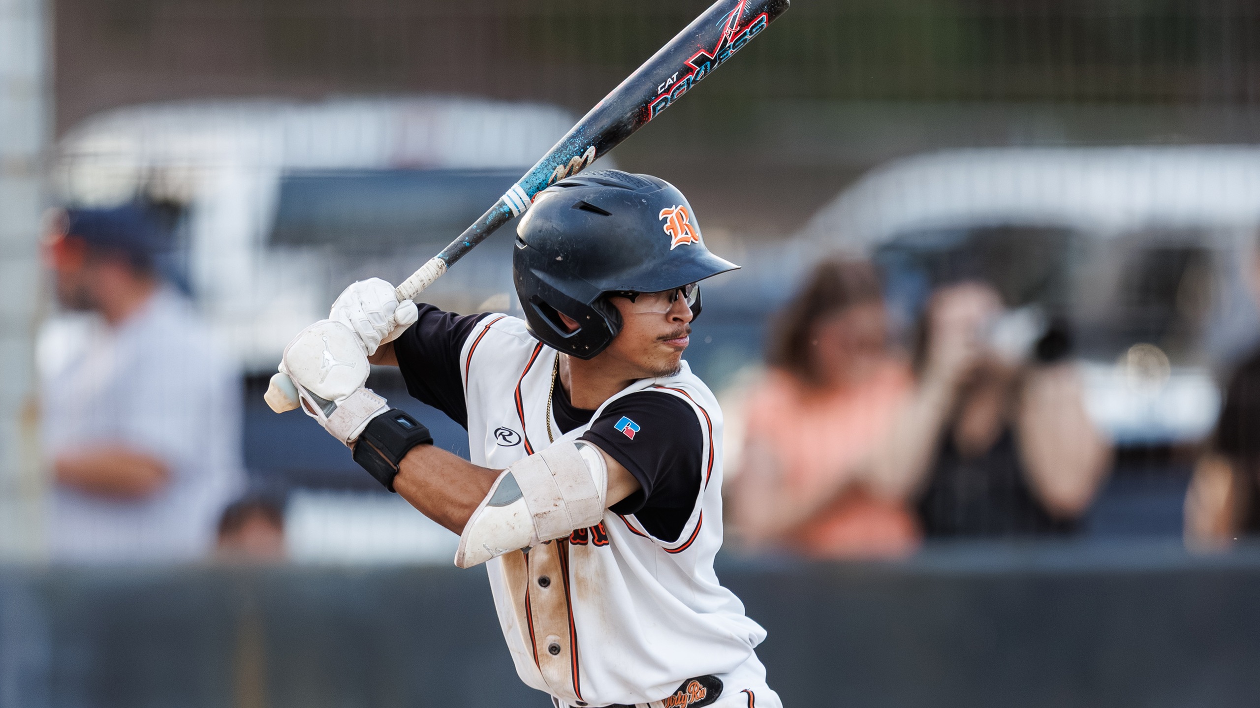 Baseball player in uniform and helmet holds bat, preparing to swing at home plate during a game, with spectators visible in the background.