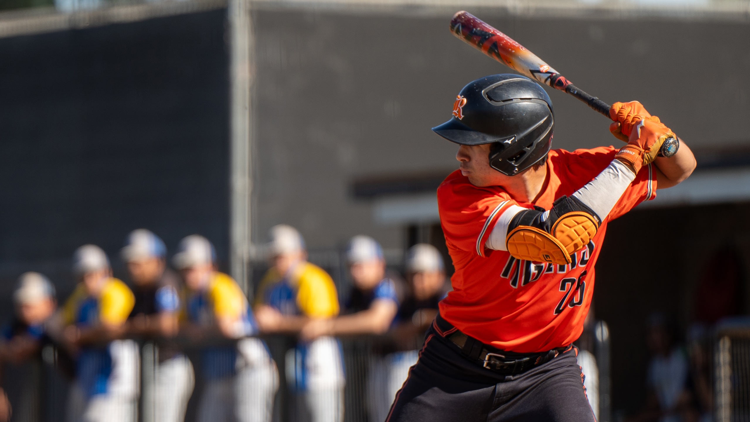 Baseball player in orange jersey and black helmet preparing to bat. Teammates in the dugout watch the game, creating a competitive atmosphere.