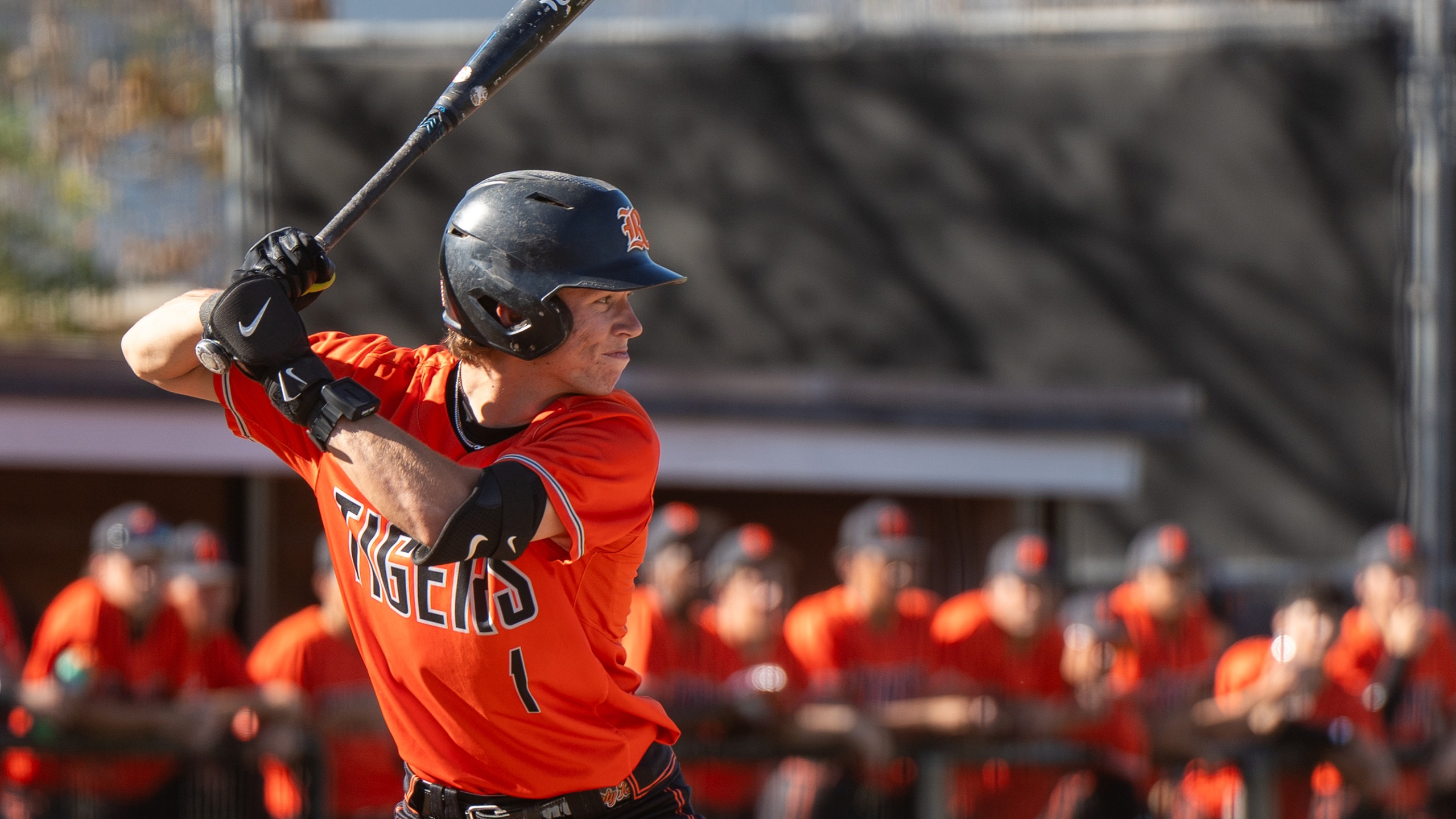 A baseball player in an orange jersey stands poised to bat, focused and ready. Behind him, teammates in matching uniforms watch intently.