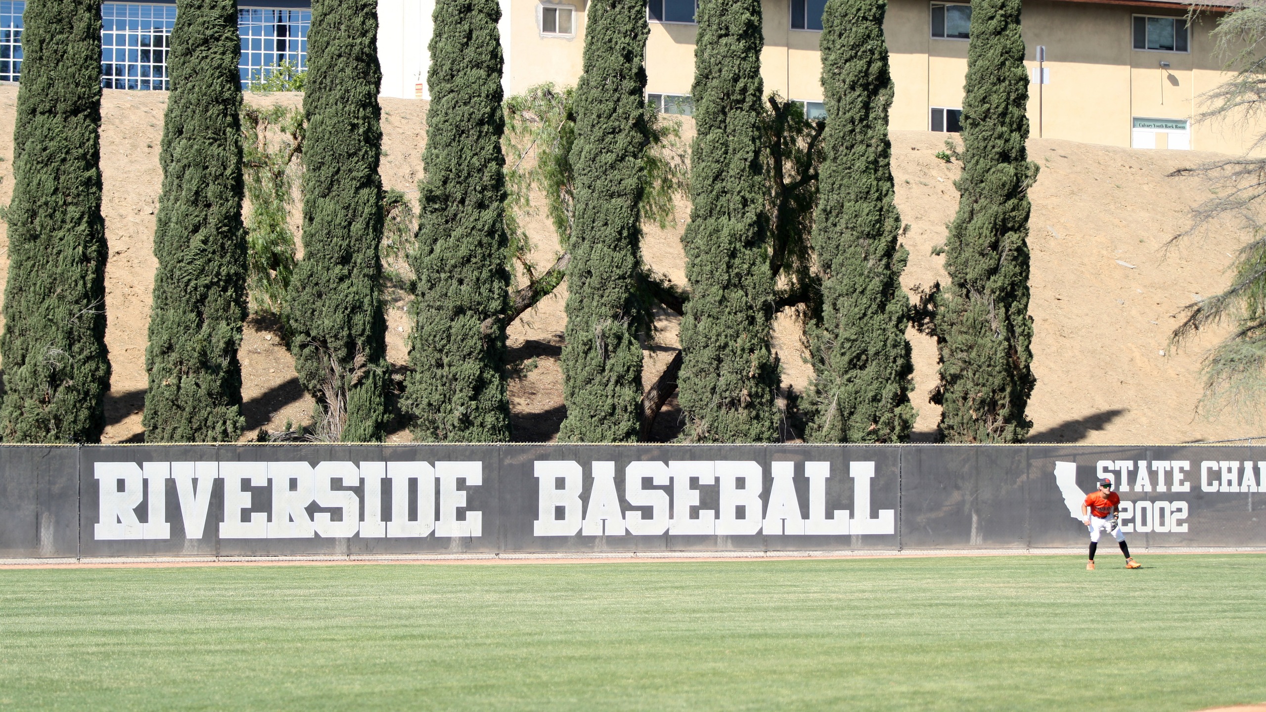 Picture of RCC baseball field with text Riverside Baseball on the fence and a man playing in the outfield.