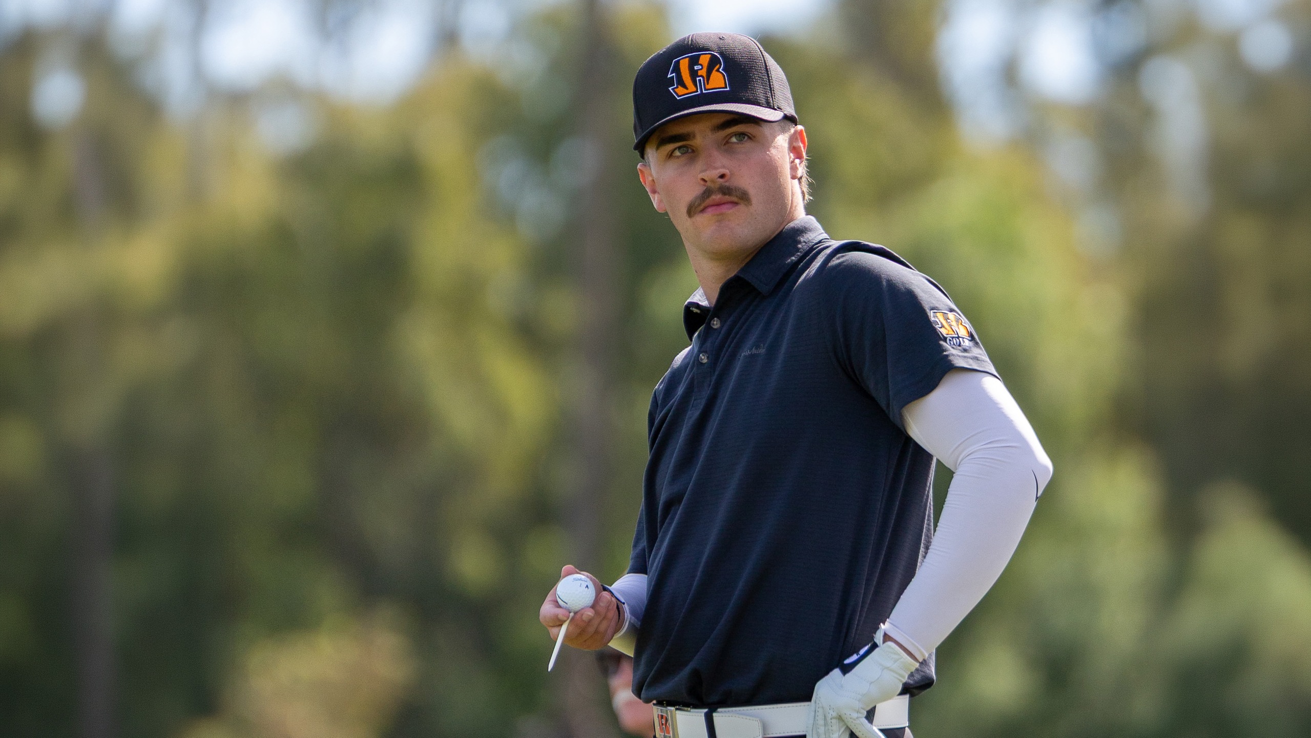 Men's golfer in a black shirt and hat stands holding a golf ball