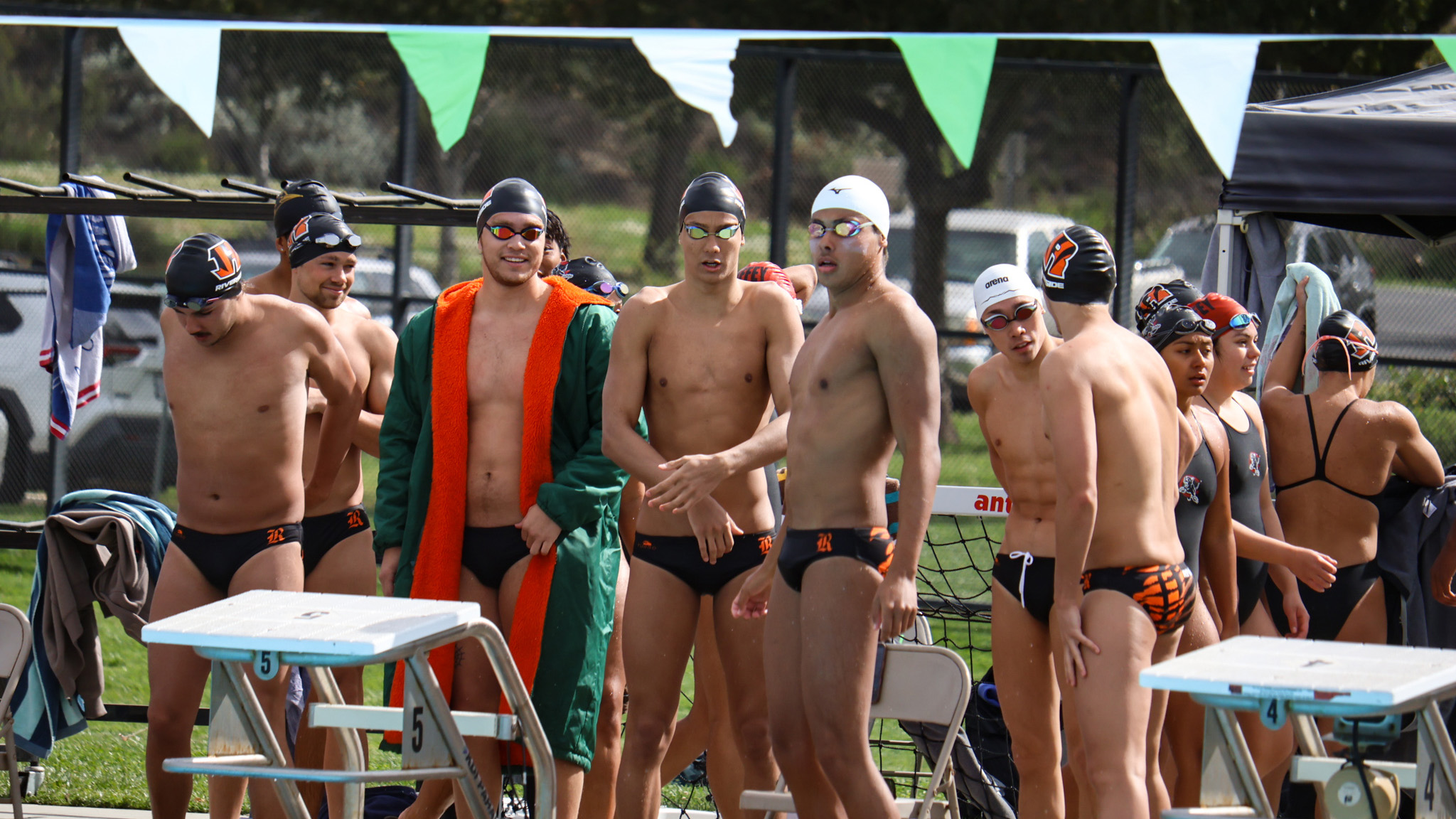 A group of young men wearing swim suits, standing together by a poolside, smiling and enjoying their time.