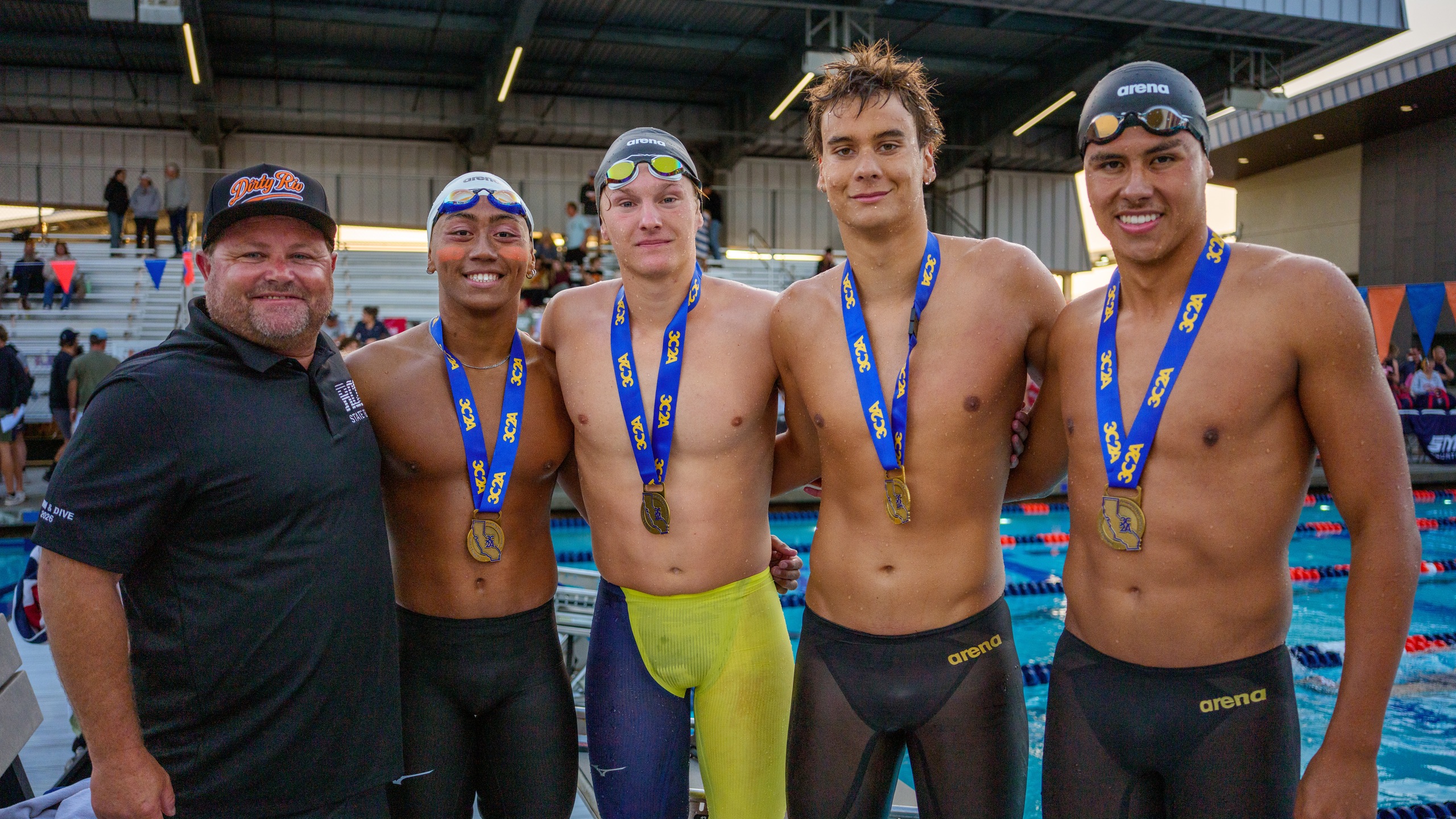 Man in a black polo poses with four male swimmers with medals around their necks.