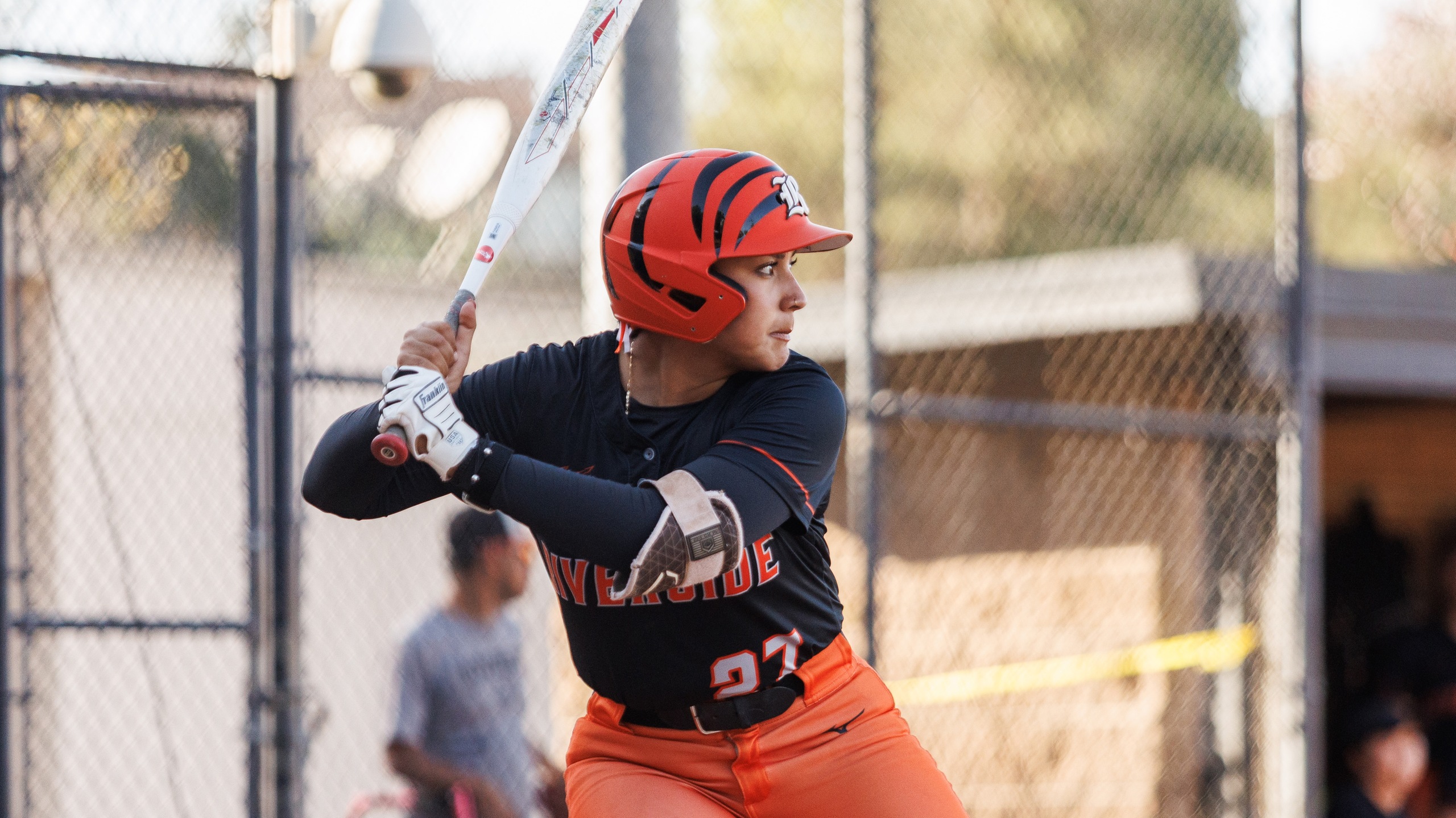 A softball player in an orange and black uniform stands ready, holding a bat in a poised batting stance.