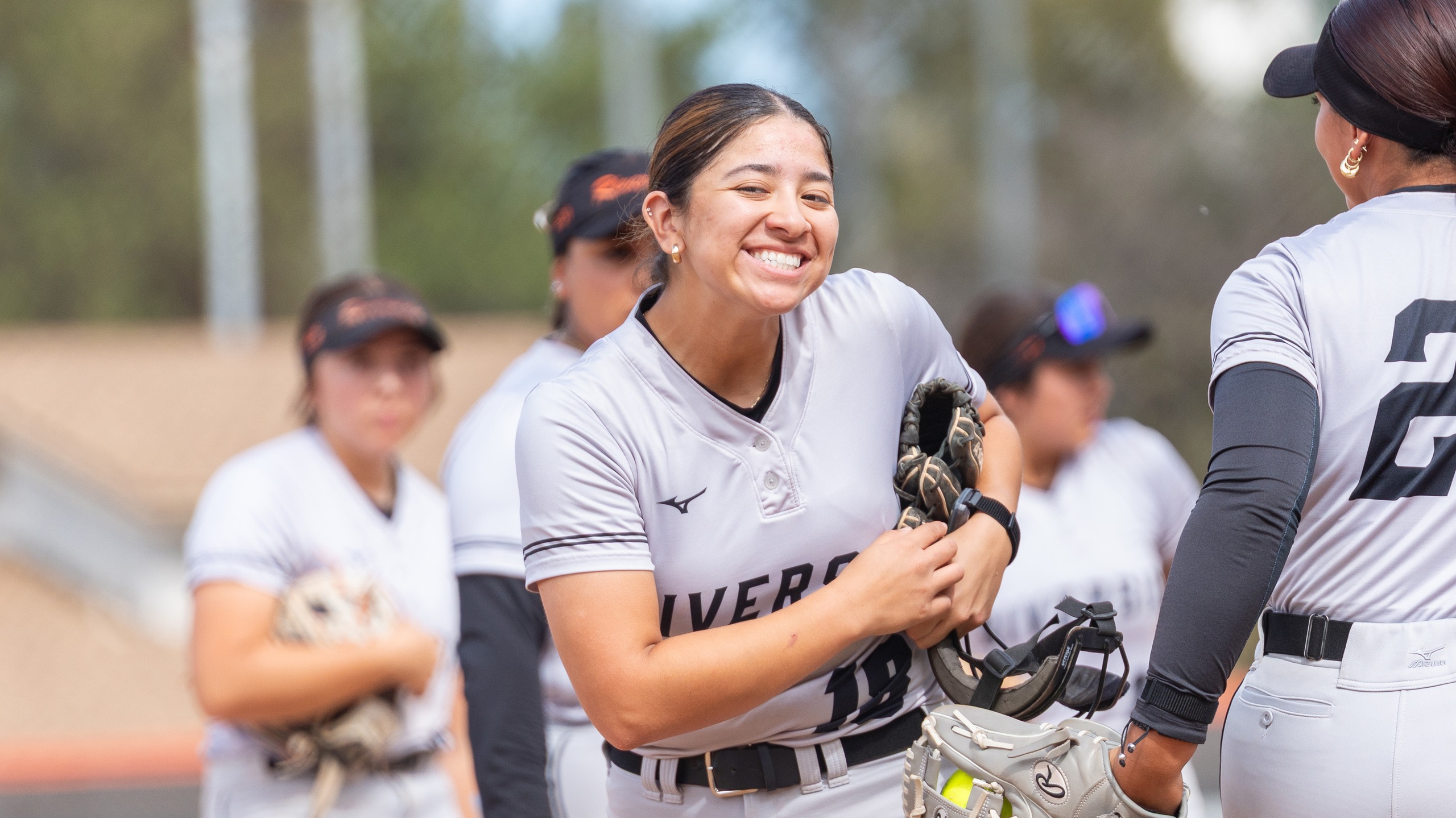 A softball player in a gray uniform joyfully smiles and high-fives a teammate. Other players, also in gray, stand in the background, conveying team spirit.