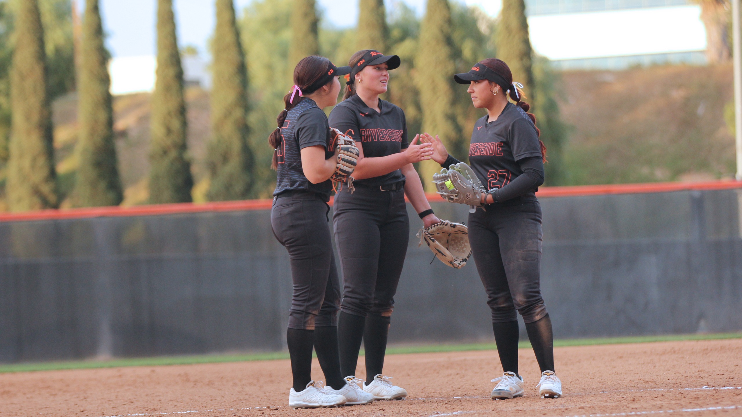Three softball players in black uniforms and caps stand on a dirt field, conversing and smiling. Background shows tall trees and a fence.