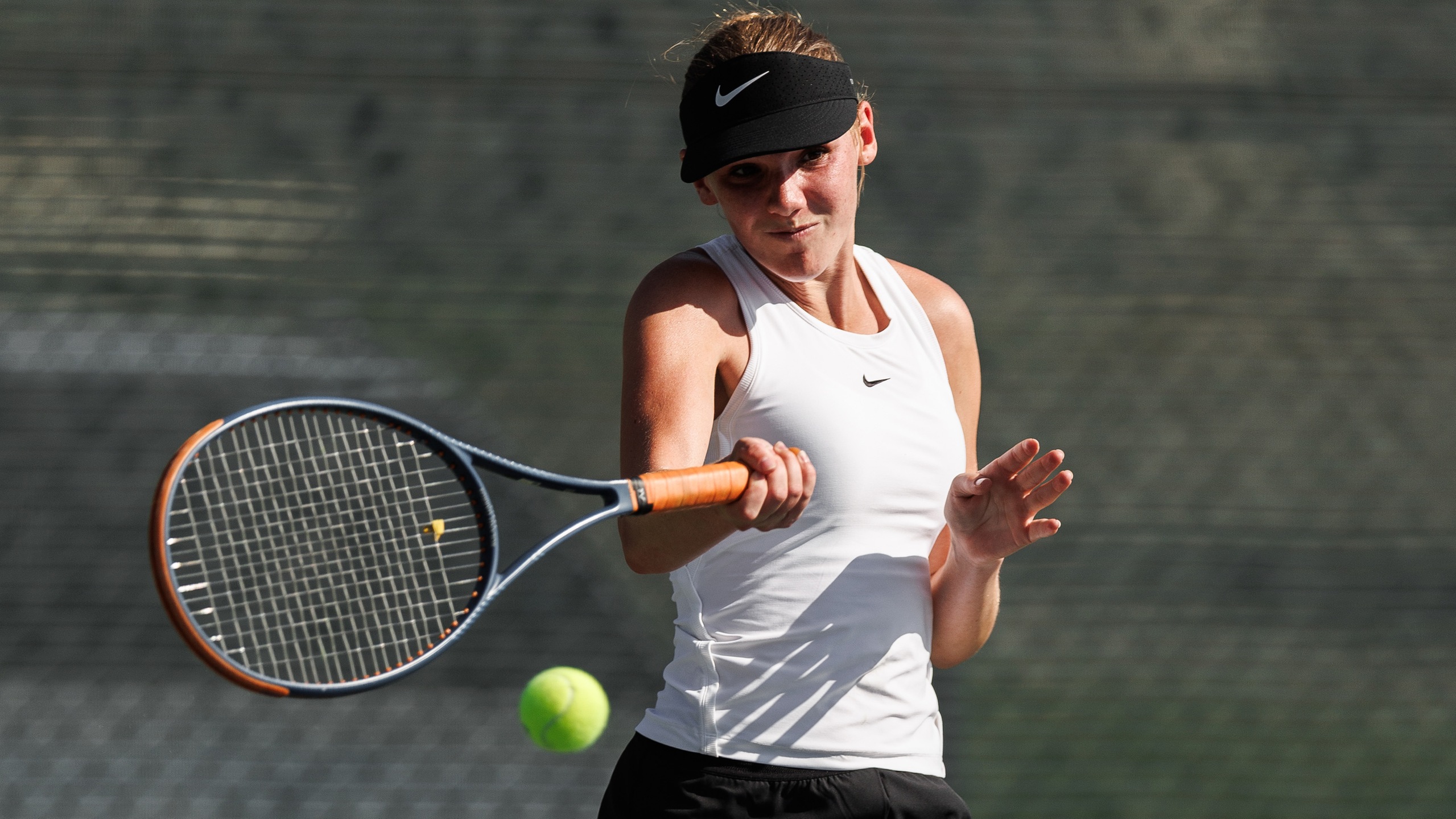 A tennis player in a white tank top and black shorts hits a forehand shot on an outdoor court.