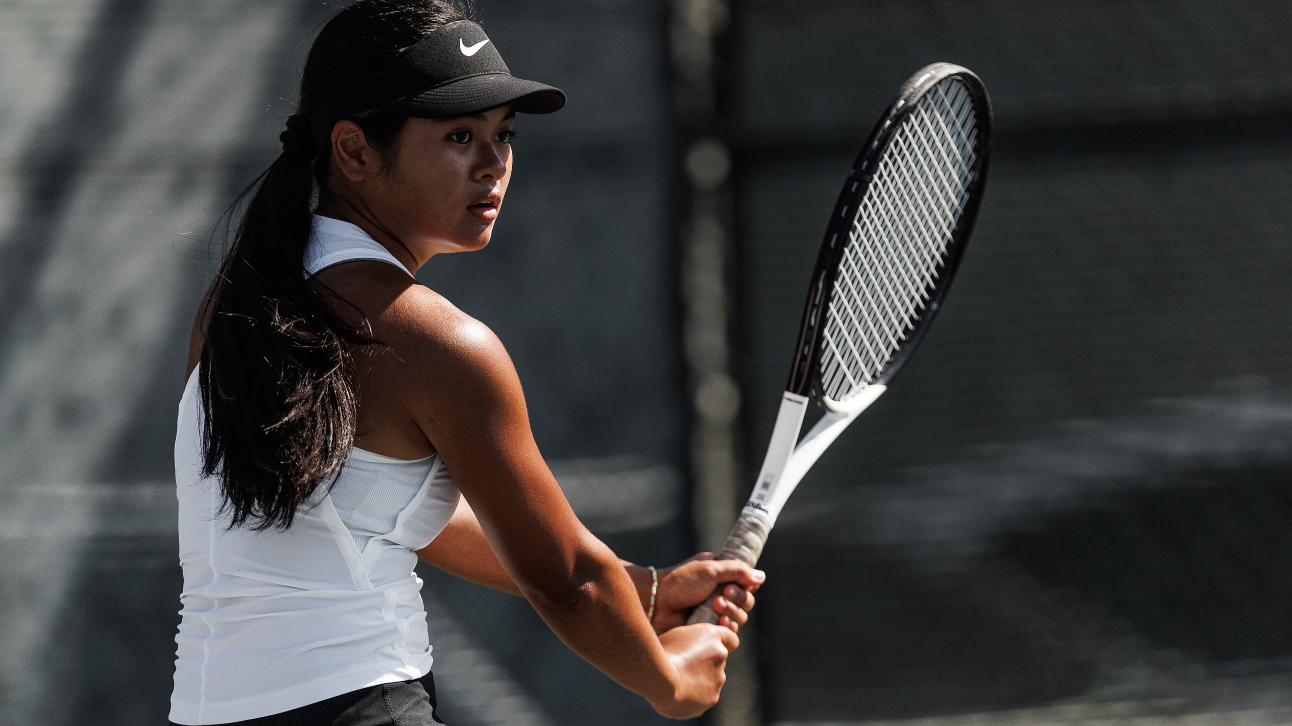 Young female tennis player in action, wearing white athletic tank top and black visor, holding a tennis racket with focused expression on outdoor hard court, sports photography, competitive tennis match, athlete mid-swing, natural sunlight, blurred background fence, high-performance training scene