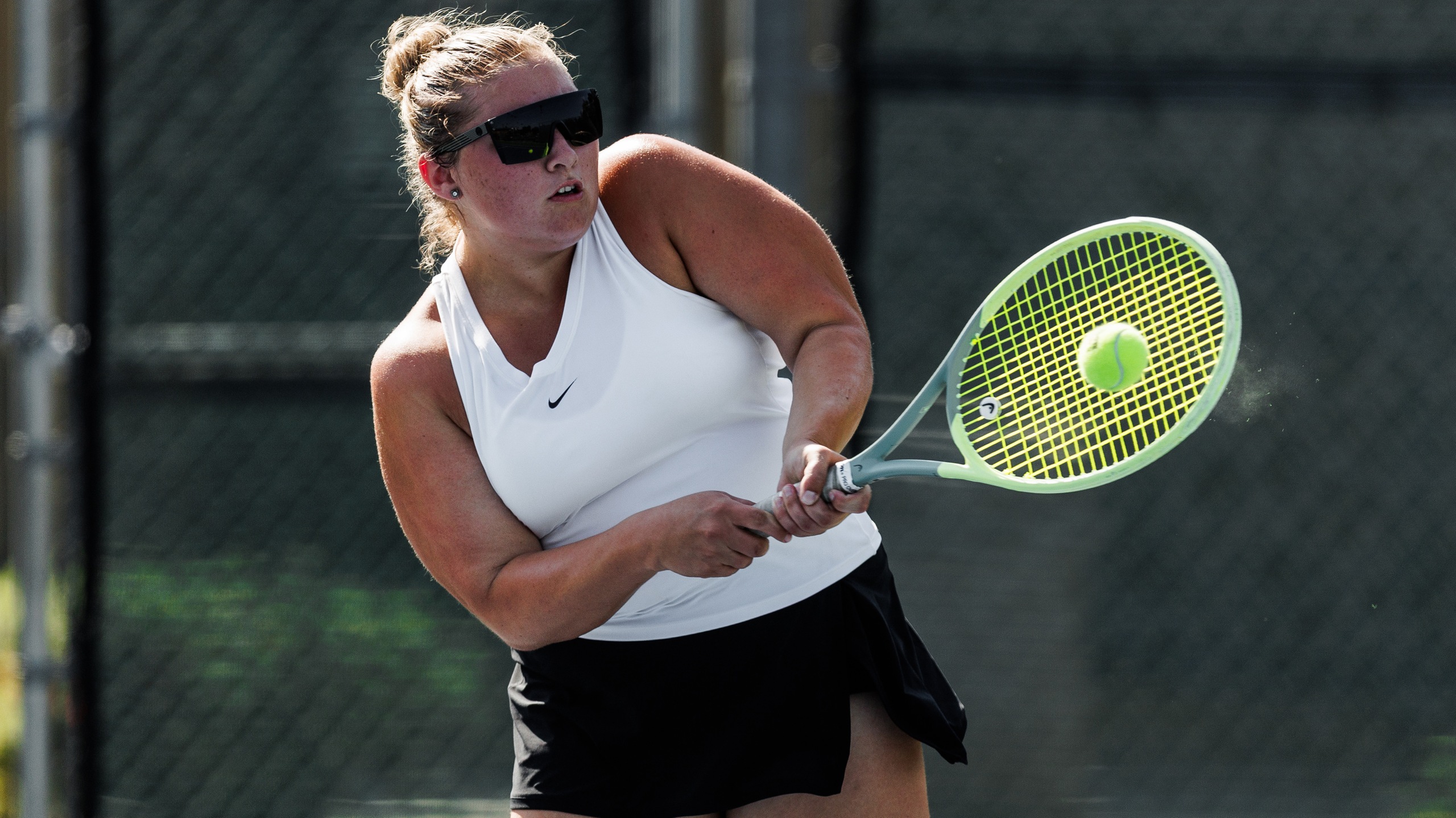 A tennis player in a white tank top and black skirt intensely focuses on hitting a ball mid-air with a vibrant racquet, conveying determination.