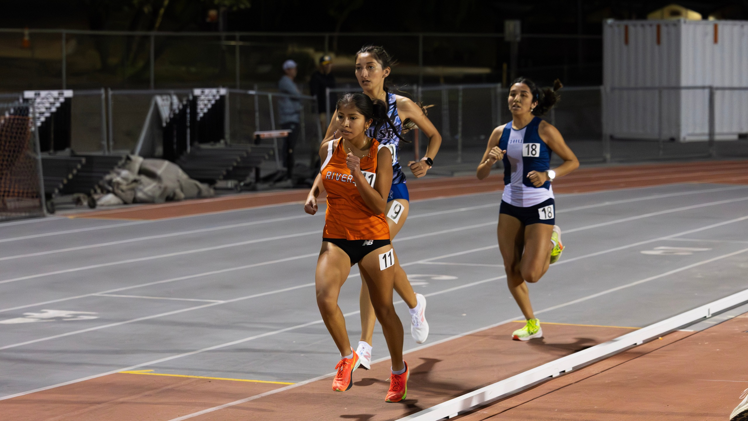 Three women running on a track at night, illuminated by stadium lights, showcasing determination and athleticism.